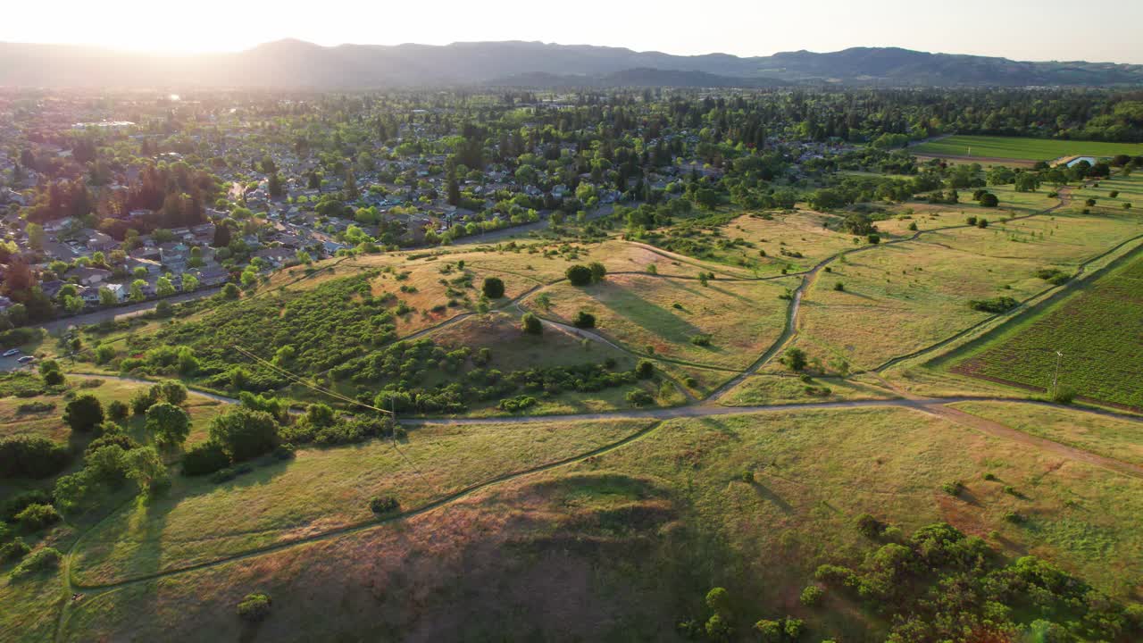 Napa Valley, California. Slow aerial wide pull-in revealing lush, vibrant green rolling hills along a hiking trail as the sun rises behind the valley's mountains, casting beams over the town below.