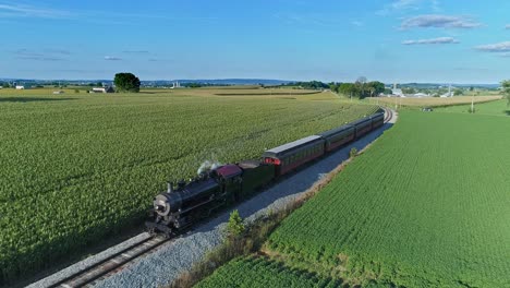 una vista aérea de un antiguo tren de pasajeros de vapor que sopla humo y vapor que viaja a través de fértiles campos de maíz en un hermoso día soleado de verano