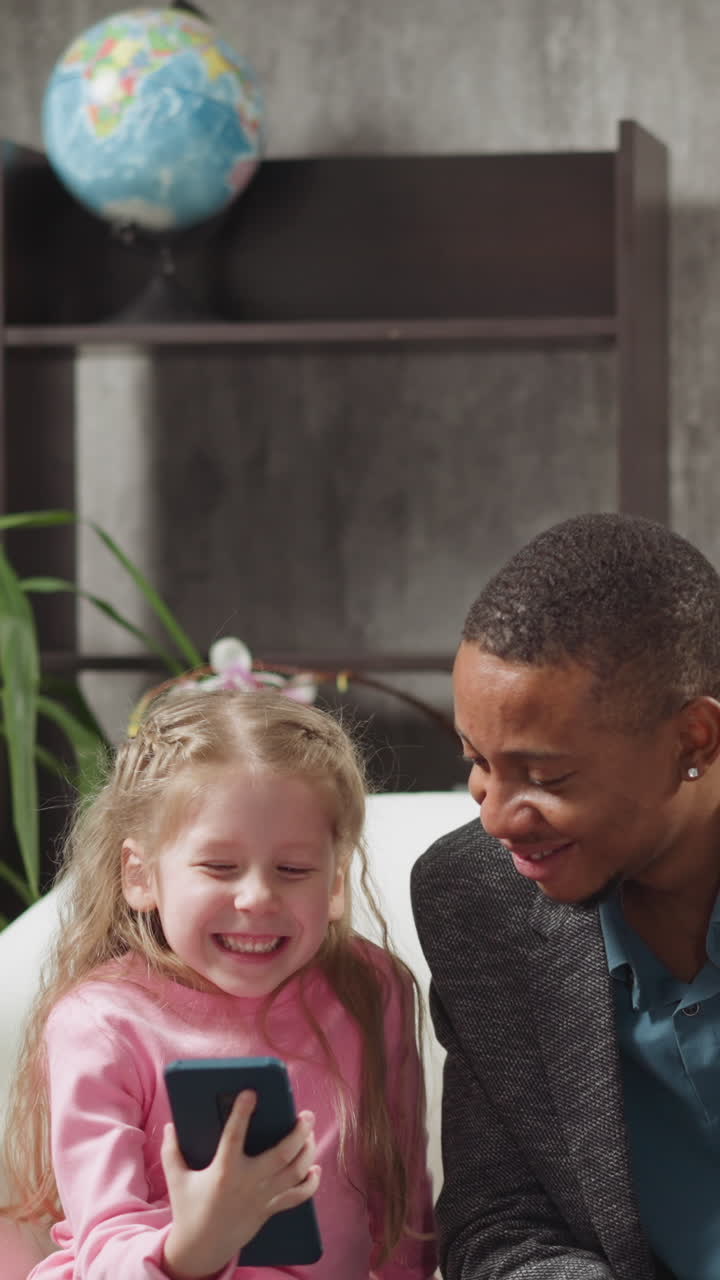 Smiling little girl watches funny videos via mobile phone with brother and African-American man teacher sitting on sofa in living room slow motion