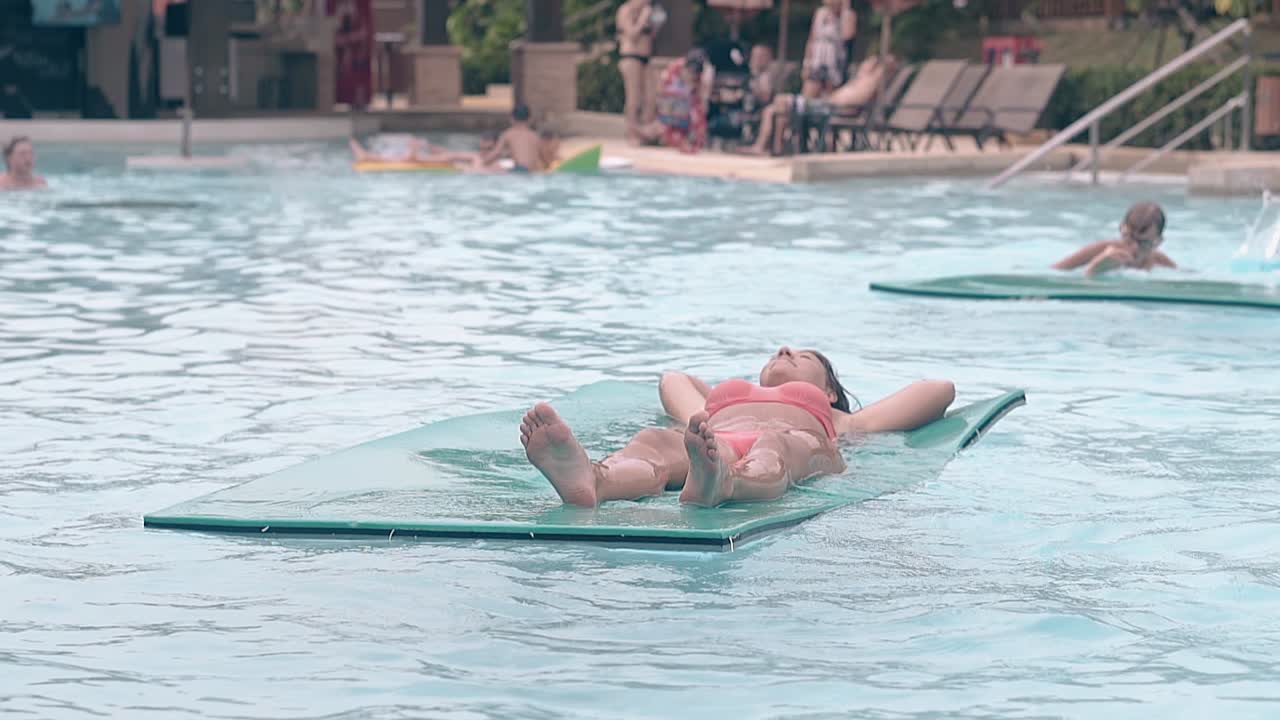 젊은 여자가 호텔 수영장에서 수영 매트에 누워 서서히 움직인다.