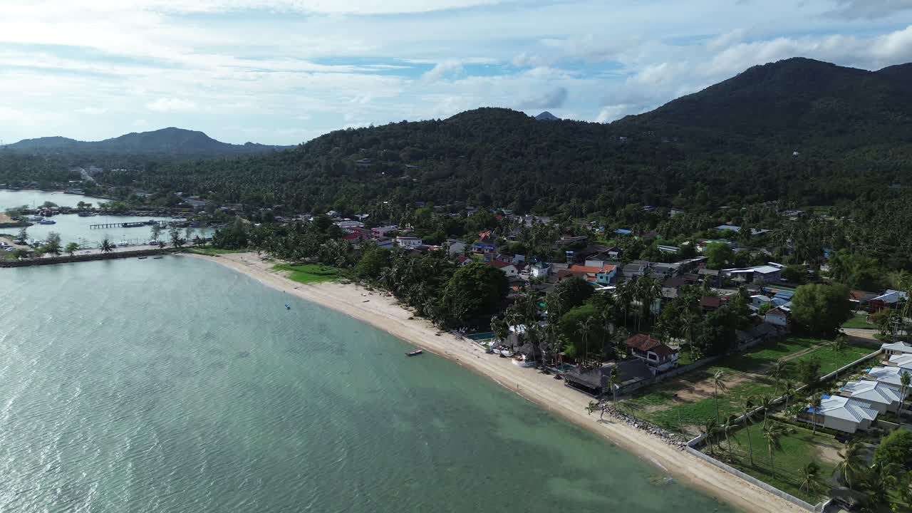 Aerial drone footage showing Baan Tai Pier and the surrounding coastal town on Koh Phangan island, Surat Thani, Thailand, with tropical hills, beach shoreline, and blue sky
