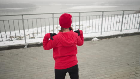 back view of woman in red hoodie and beanie performing arm stretches by lifting hands to shoulders and turning arms outdoors, misty atmosphere with river and iron railing in background