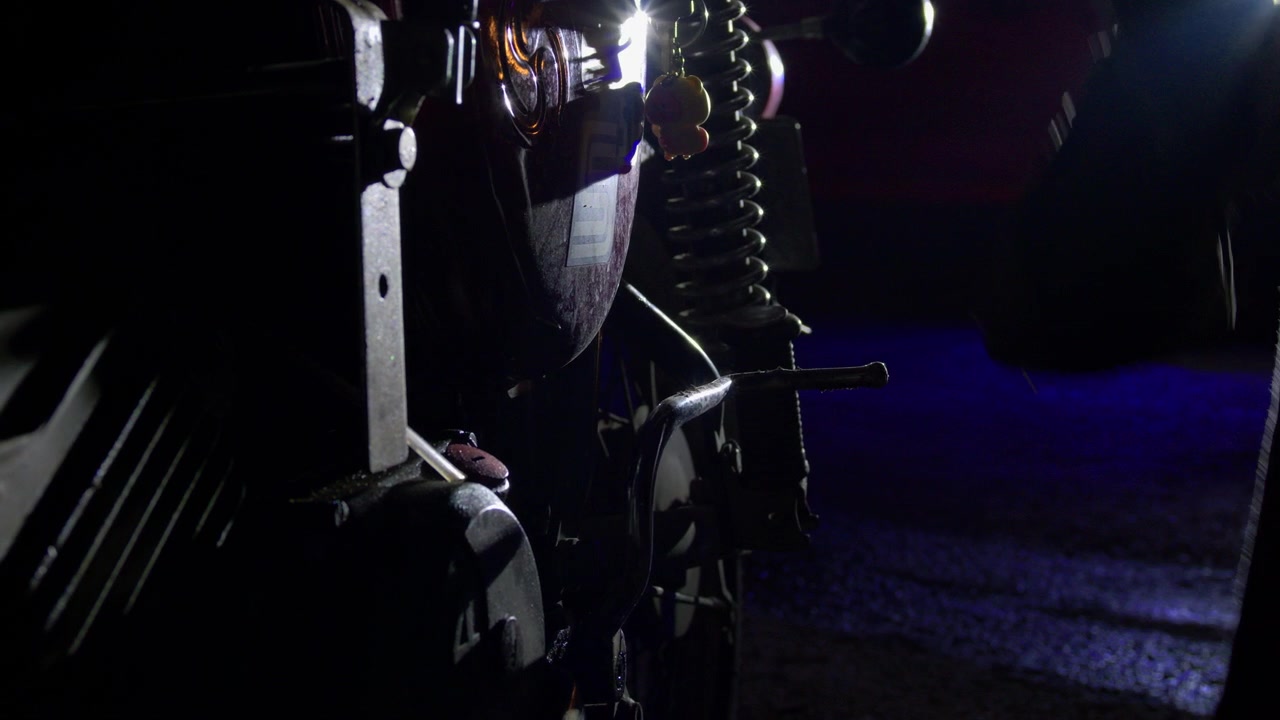 Primo piano di una motocicletta in manutenzione di notte, con sospensioni, ruota e parti meccaniche illuminate da luce blu in un ambiente stradale buio, creando una scena di manutenzione cinematografica