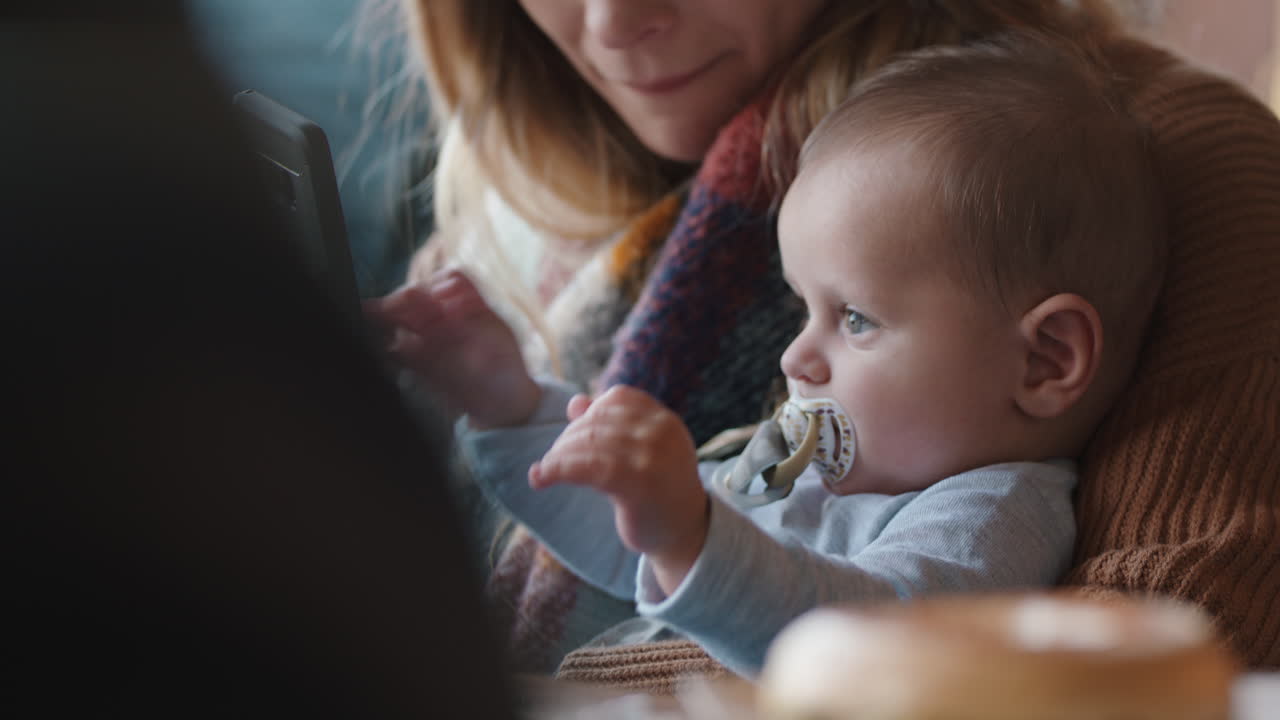 아기를 데리고 카페에 있는 젊은 엄마는 분주한 레스토랑에서 스마트폰으로 커피를 마시며 모성을 즐기고 있습니다.