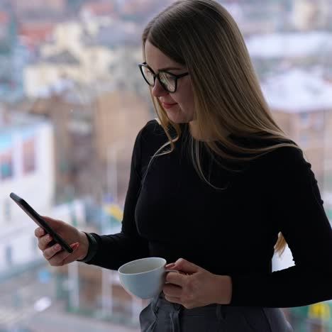 beautiful female with cup of coffee thoughtfully browsing smartphone while standing by the window