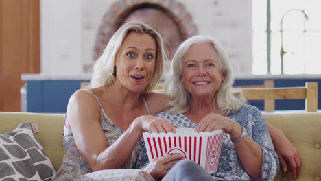 madre con hija adulta comiendo palomitas de maíz viendo una película en el sofá en casa juntos