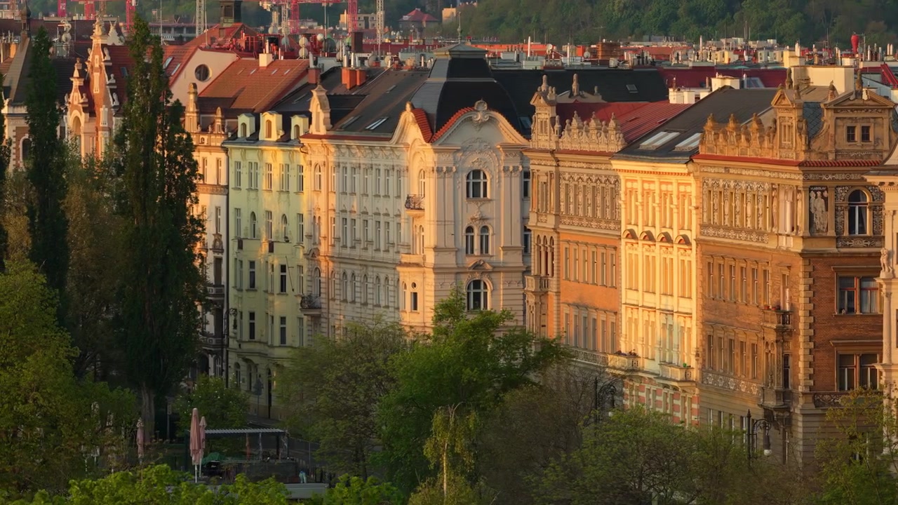 Vista aerea con drone di facciate residenziali ornate in stile Art Nouveau e neobarocco, illuminate dalla calda luce dell'ora d'oro, con le chiome degli alberi in primo piano, Praga, Repubblica Ceca.