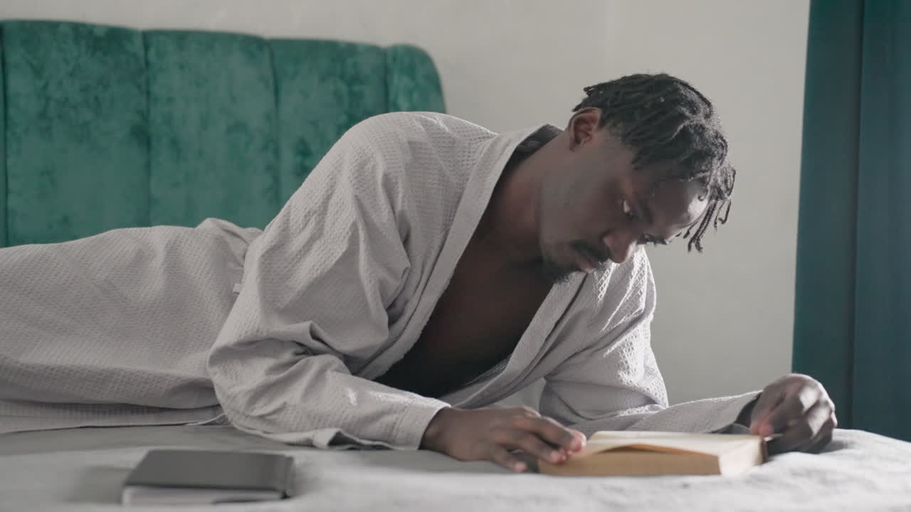 Guy in white bathrobe laying on bed, focusing on reading a book, flipping pages with one hand, green velvet headboard in background, soft natural light illuminating relaxed atmosphere