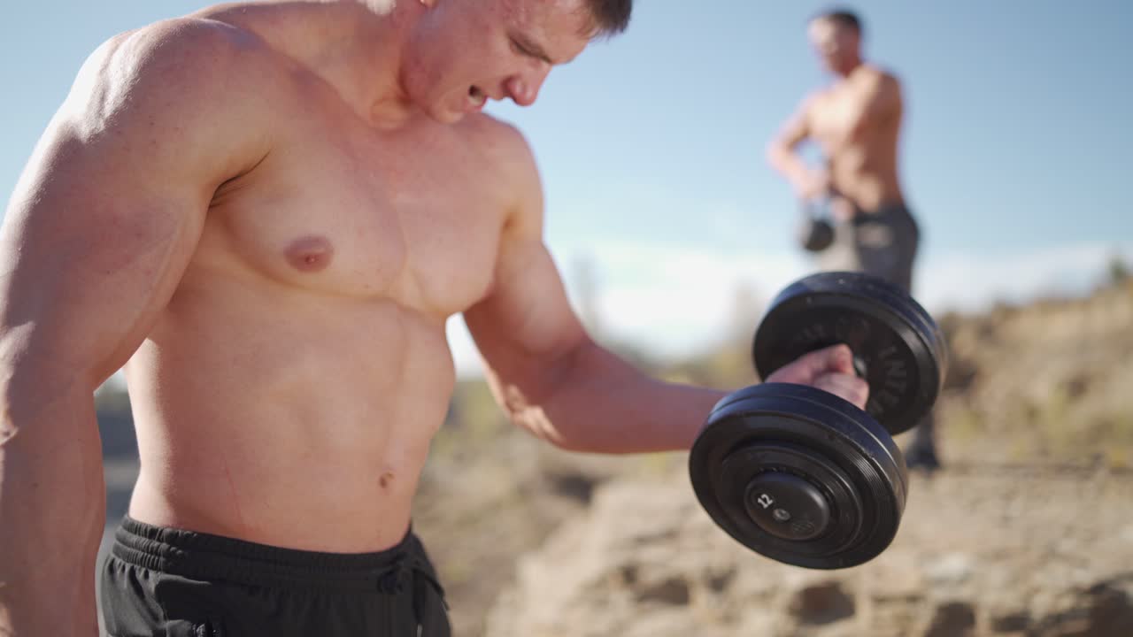 Healthy bodybuilder with heavy dumbbells. Muscular sportsman lifting metal dumbbells outdoors. Physical workout in nature. Close-up.