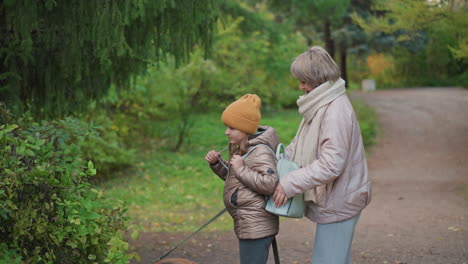 mom adjusts daughter bag strap while guiding dog walk home on leafy trail, bundled in jackets and scarf under soft autumn light, intimate family moment showcasing care and togetherness