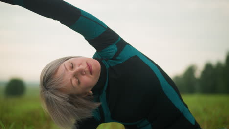 middle-aged woman lying on one side, practicing side bend yoga pose with arm extended, eyes closed, in a vast grassy field under cloudy skies, with slightly blurred trees in the distance