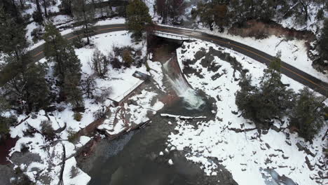 Aerial-pullback-of-mountain-stream-waterfall-flowing-into-freezing-lake-under-overpass-as-vehicle-drives-over-on-road