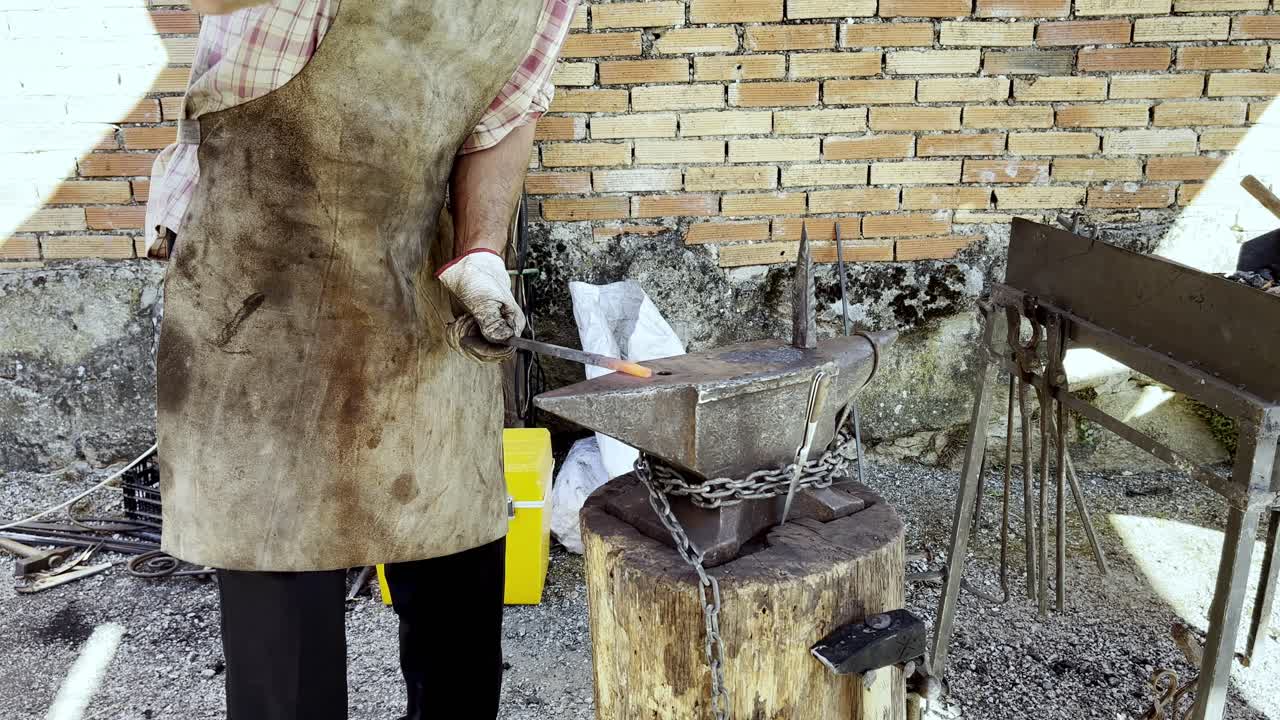 el herrero gira y martilla una pieza de metal caliente rojo contra el yunque encadenado a un tronco de madera