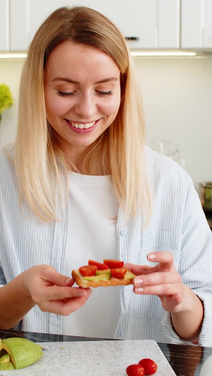 Giovane donna che mangia toast all'avocado con fette di pomodoro per una colazione sana e nutriente nella cucina di casa.