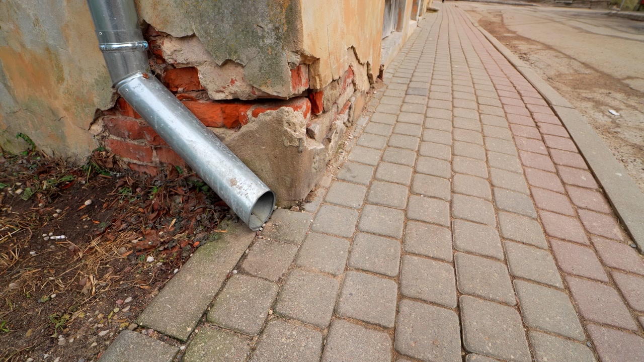 Broken downspout drains onto a sidewalk beside exposed brick and crumbling plaster, showing poor drainage and a weathered facade on an old urban building.