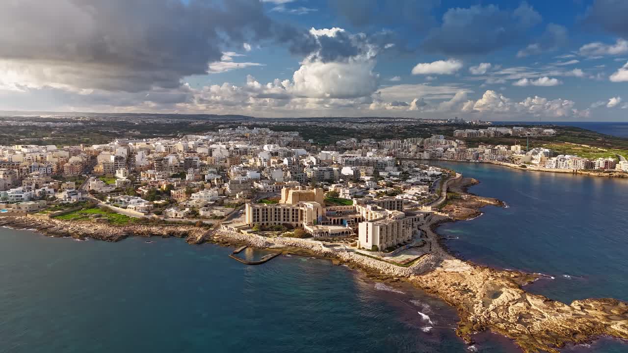 Hyperlapse orbitale con drone attorno a Marsascala, Malta, che mette in risalto il suo fascino urbano costiero, le acque mediterranee, la penisola panoramica e il vivace porto sotto un cielo azzurro nuvoloso in una soleggiata giornata invernale