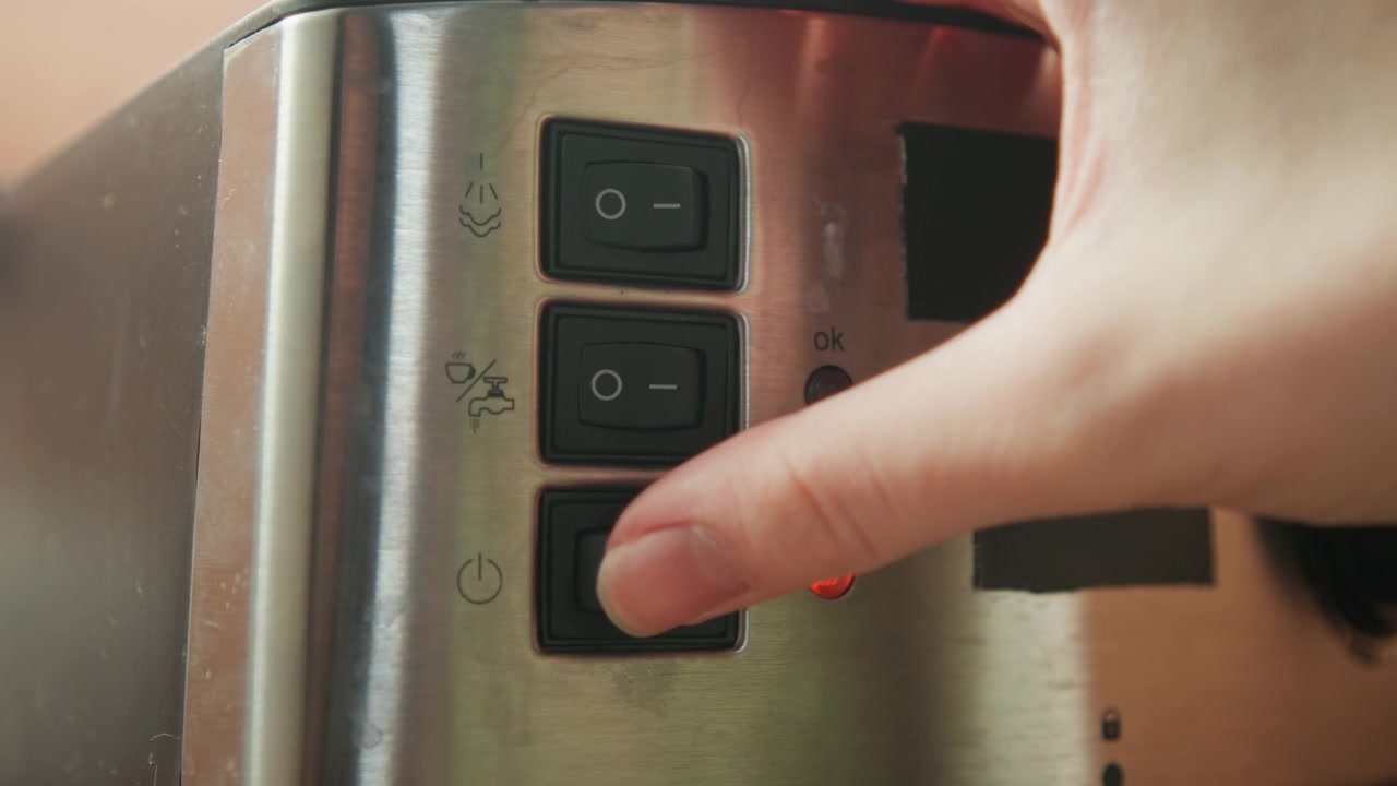 Angled close view shows fingertip pressing black rocker switch on stainless appliance control panel, with red indicator glowing, smudged brushed metal surface, taped section, and visible wear
