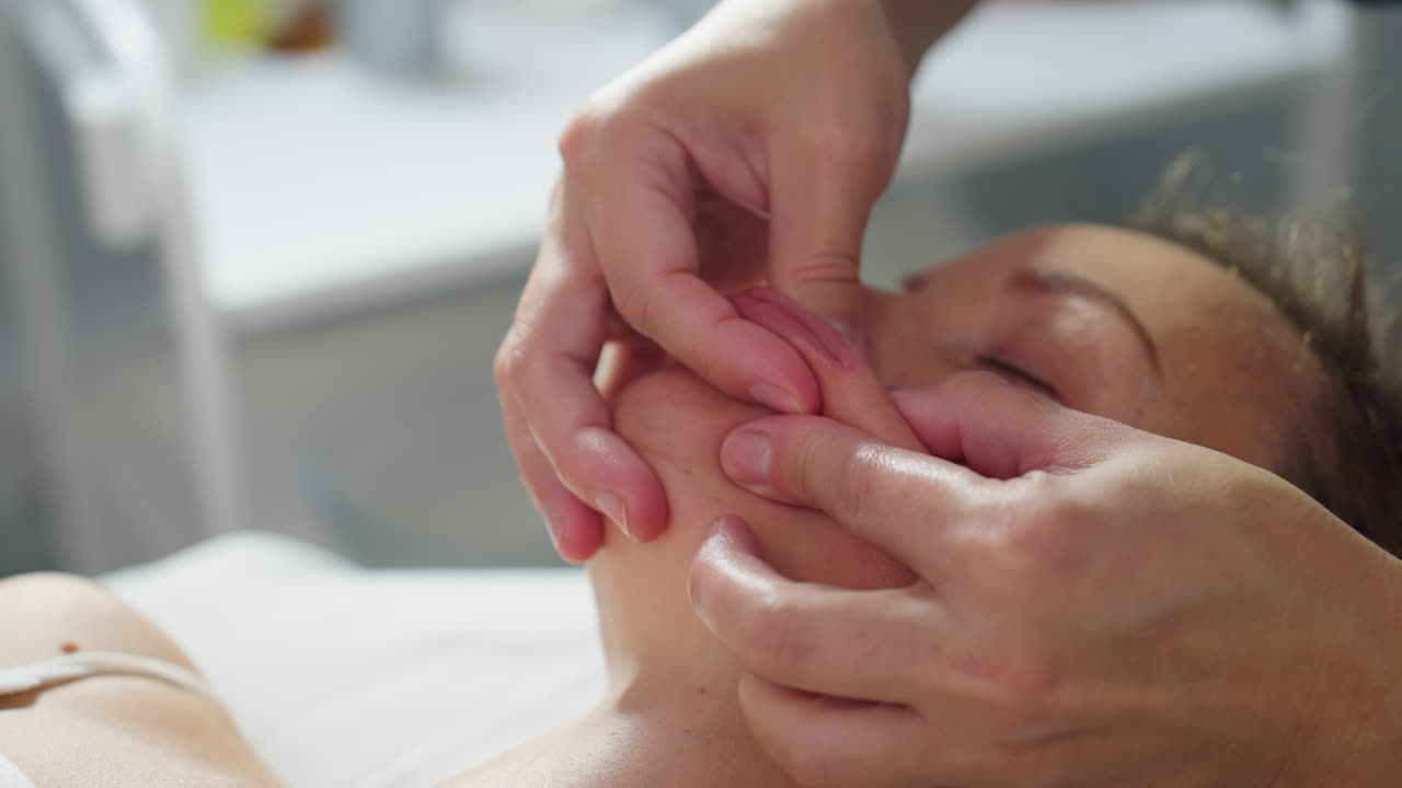 Close up of relaxed client lying on massage bed with eyes closed as therapist gently massages lip and cheek area using cream and soft strokes for soothing facial treatment and rejuvenating skin glow