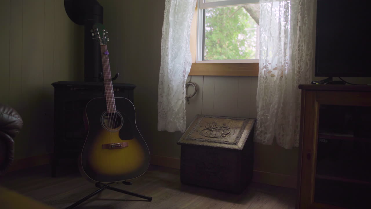 A guitar sits on a stand by an open window as the wind blows the curtains around. Establishing shot