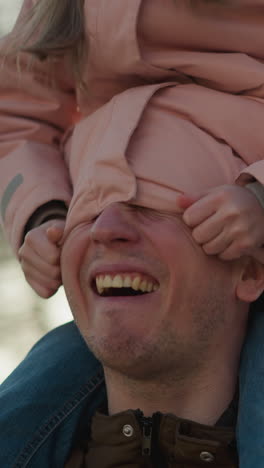 un momento alegre cuando una niña con una gorra y una chaqueta rosadas se sienta en el cuello de su padre, metiendo juguetonamente su cabeza dentro de su chaqueta. ambos están riendo, capturando una interacción conmovedora y lúdica