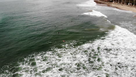 Aerial-drone-shot-on-a-cloudy-day-over-a-group-of-surfers-in-wetsuits-catching-and-surfing-waves-in-the-Pacific-Ocean-off-the-coast-of-Ventura,-California