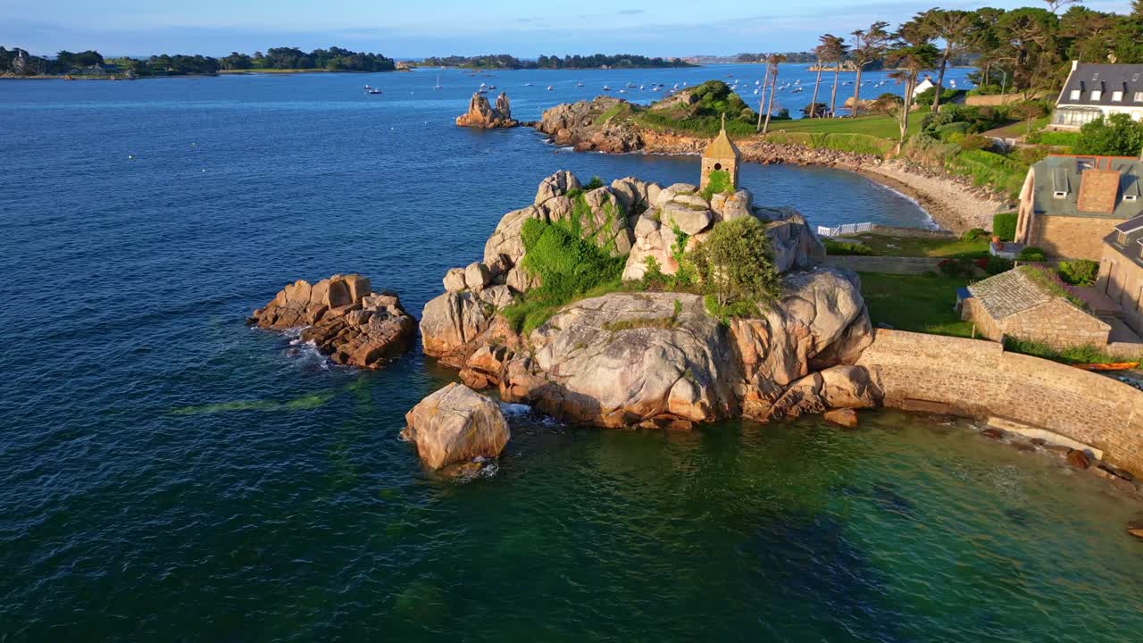 Approaching drone movement to the rocky coastal formation with The Sentinel Oratory building over turquoise waters and seaside cottages at Port Blanc, Côtes-d'Armor, Brittany, France.