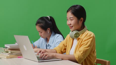 close up of asian woman student typing on a laptop and smiling to camera while sitting with her friend studying on a table in the green screen background classroom