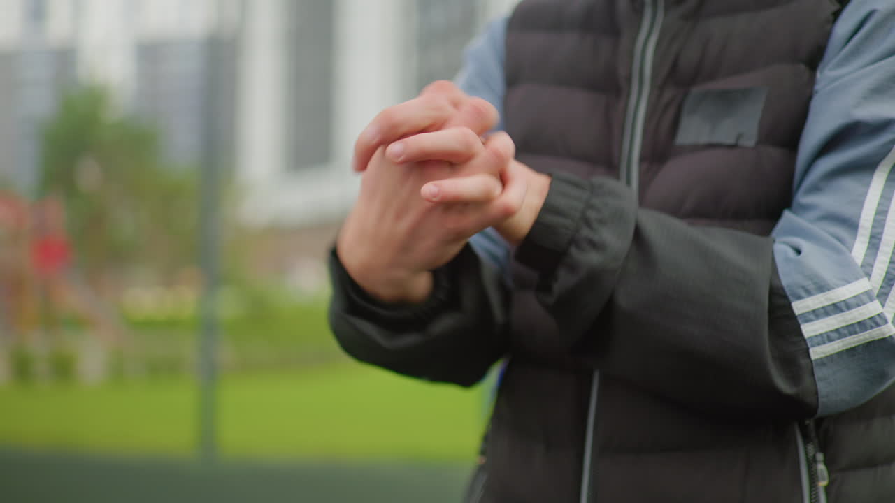 Person, der varmer fingrene i en varm jakke i en park i byen, tæt på. Vatteret ærme med hvide striber er synligt. Subtil gnidningsbevægelse antyder pre-workout rutine, køligt overskyet lys.