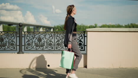 lady in grey clothing walking near riverfront holding shopping bag, passing a decorative metal fence, background shows a blurred bridge and greenery under a clear sky, with a shadow cast on her face