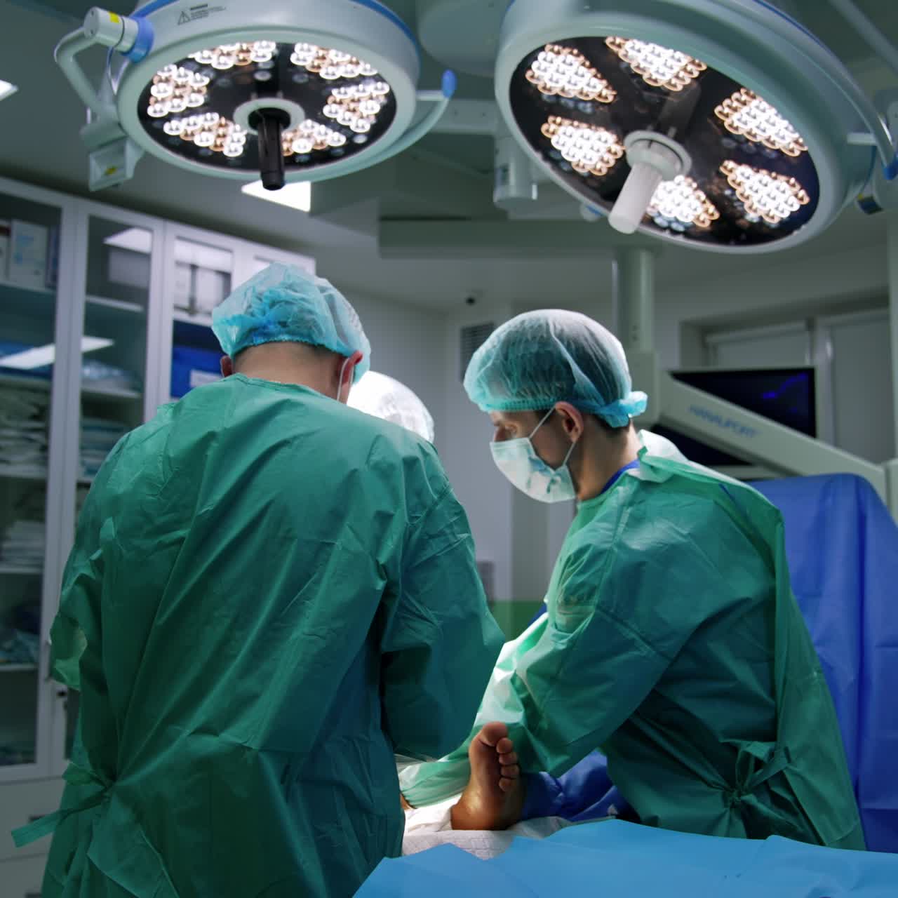 Un ayudante apoya el pie del paciente operado en un moderno quirófano. Dos lámparas redondas iluminan al equipo médico