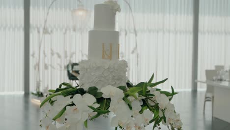 three tier white wedding cake with floral details and gold initials, displayed on an orchid arrangement