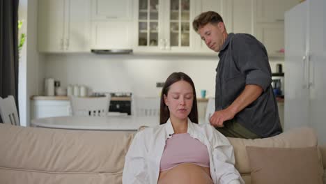 a middle-aged man approaches his pregnant wife and sits on the sofa near her while showing his concern for his wife at home