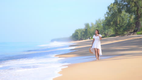 Eine-Junge-Fitte-Frau-In-Einem-Weißen-Sommerkleid-Geht-Am-Strand-Entlang,-Während-Die-Wellen-Hereinrollen