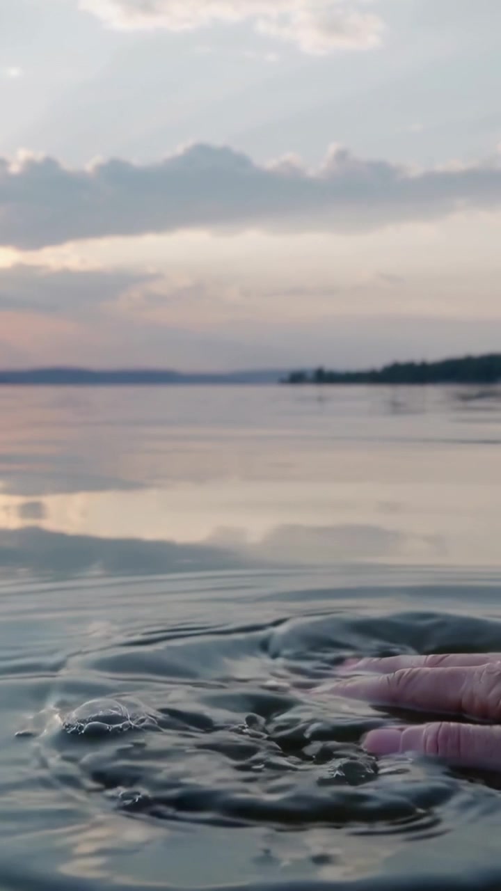 Video verticale: Mano nuda che entra disturbando la superficie del lago al tramonto, formando increspature e bolle.