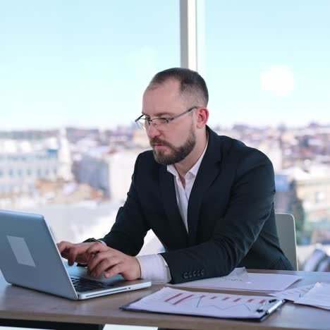 Serious business worker on office window backdrop. Middle aged businessman in glasses sitting at desk in his workplace. Entrepreneur searches solution of business issues