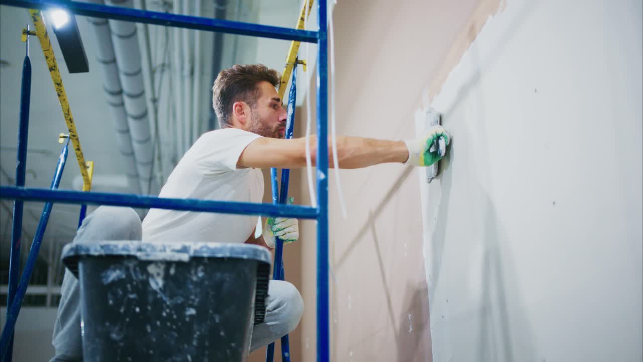 A Skilled Worker Carefully Applies Finishing Touches to a Wall Using a Trowel While Balancing on a Scaffolding Structure in a Modern Interior Space