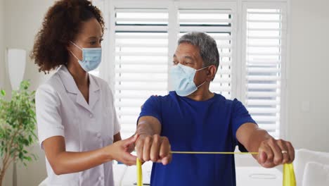 Mixed-race-female-physiotherapist-wearing-mask-helping-senior-exercise-using-exercise-band