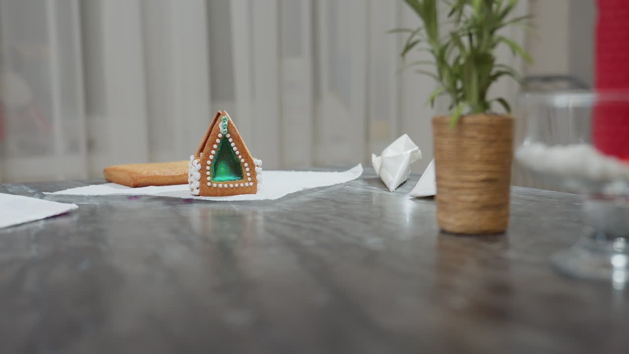 una casa de pan de jengibre bellamente decorada con una ventana de azúcar verde esmeralda se sienta en una mesa texturizada, rodeada de dos bolsas de azúcar de hielo y elementos de cocina borrosos en el fondo