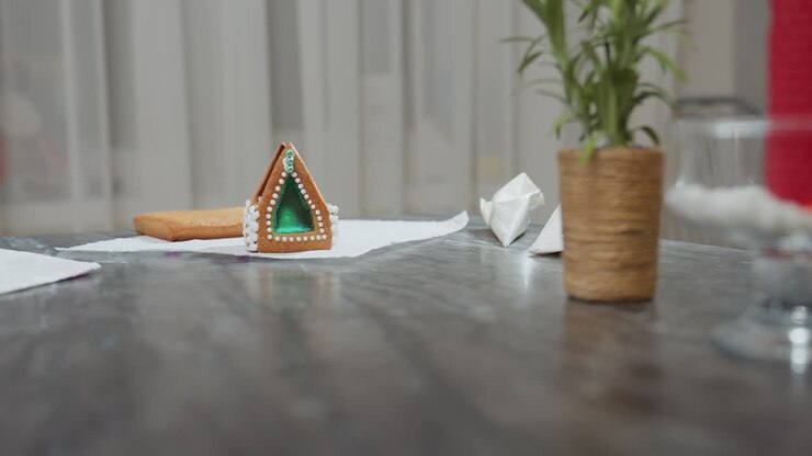 Una casa di pan di zenzero splendidamente decorata con una finestra di zucchero verde smeraldo è appoggiata su un tavolo texturizzato, circondata da due sacchetti di zucchero a velo ed elementi sfocati della cucina sullo sfondo