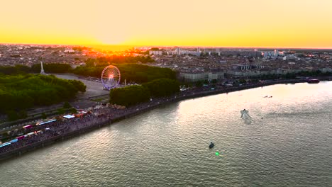 Feria-Del-Vino-Con-Noria-Iluminada-Al-Atardecer-Con-Multitudes-En-Burdeos,-Francia,-Con-Río-Garona-Y-Barcos,-Toma-Aérea-De-Paso-Elevado