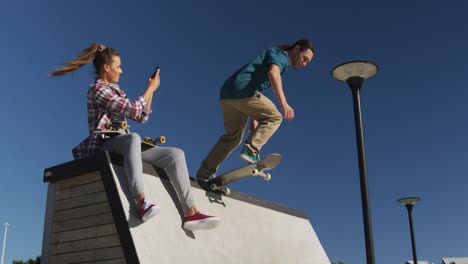 happy caucasian woman taking photo of her male friend skateboarding