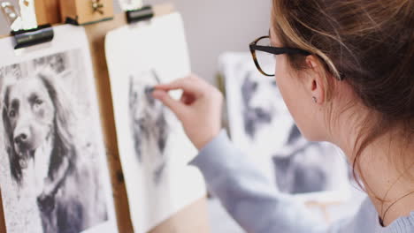 Female-Teenage-Artist-Sitting-At-Easel-Drawing-Picture-Of-Dog-From-Photograph-In-Charcoal