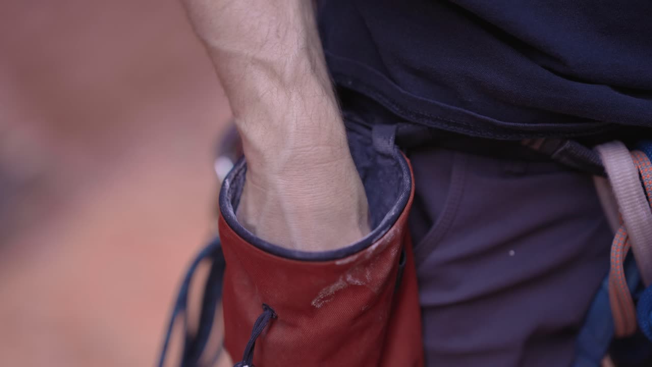 A close-up detail shot of a rock climber dipping their hand into a chalk bag, applying chalk to their fingers for a better grip before starting a climb