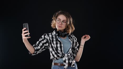 una joven linda con camisa a cuadros y auriculares en el cuello preparándose, mirando a su teléfono inteligente y tomando una selfie aislada en un fondo negro en el estudio. cara de mueca, mostrando la lengua
