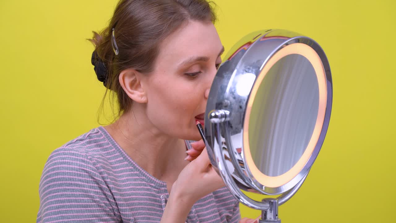 평범한 줄무 셔츠를 입은 젊은 성인 아름다운 여성, 은 연필로 입술 모양을 윤, 금속 프레임의 두 표면을 가진 둥근 거울을보고. 밝은 노란색 배경, 스튜디오.