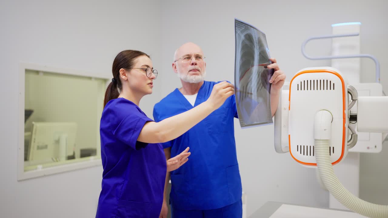 A confident brunette girl with glasses in a blue uniform communicates together and consults with a confident man doctor with gray hair while summing up the results of fluorography and confirming the diagnosis in a modern clinic