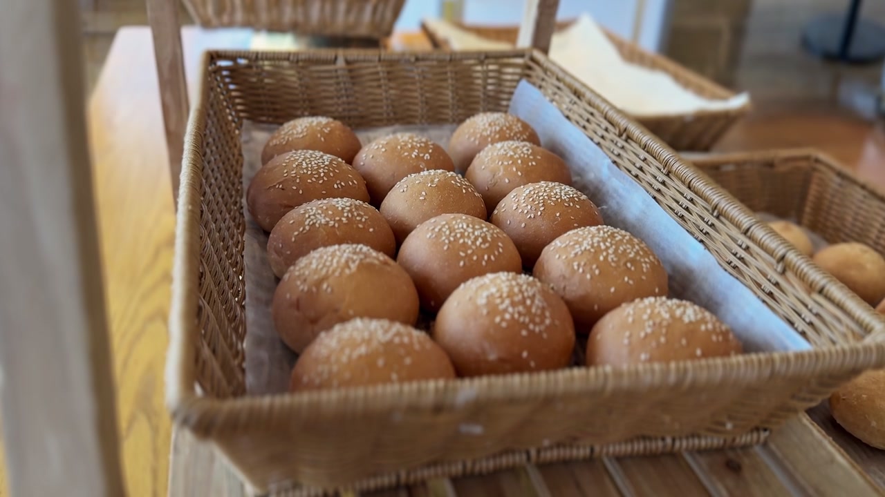 Close-up of fresh baked bread buns with sesame seeds inside a woven wicker basket lined with parchment paper. Bright indoor daylight highlights the bakery food display with a shallow depth field.