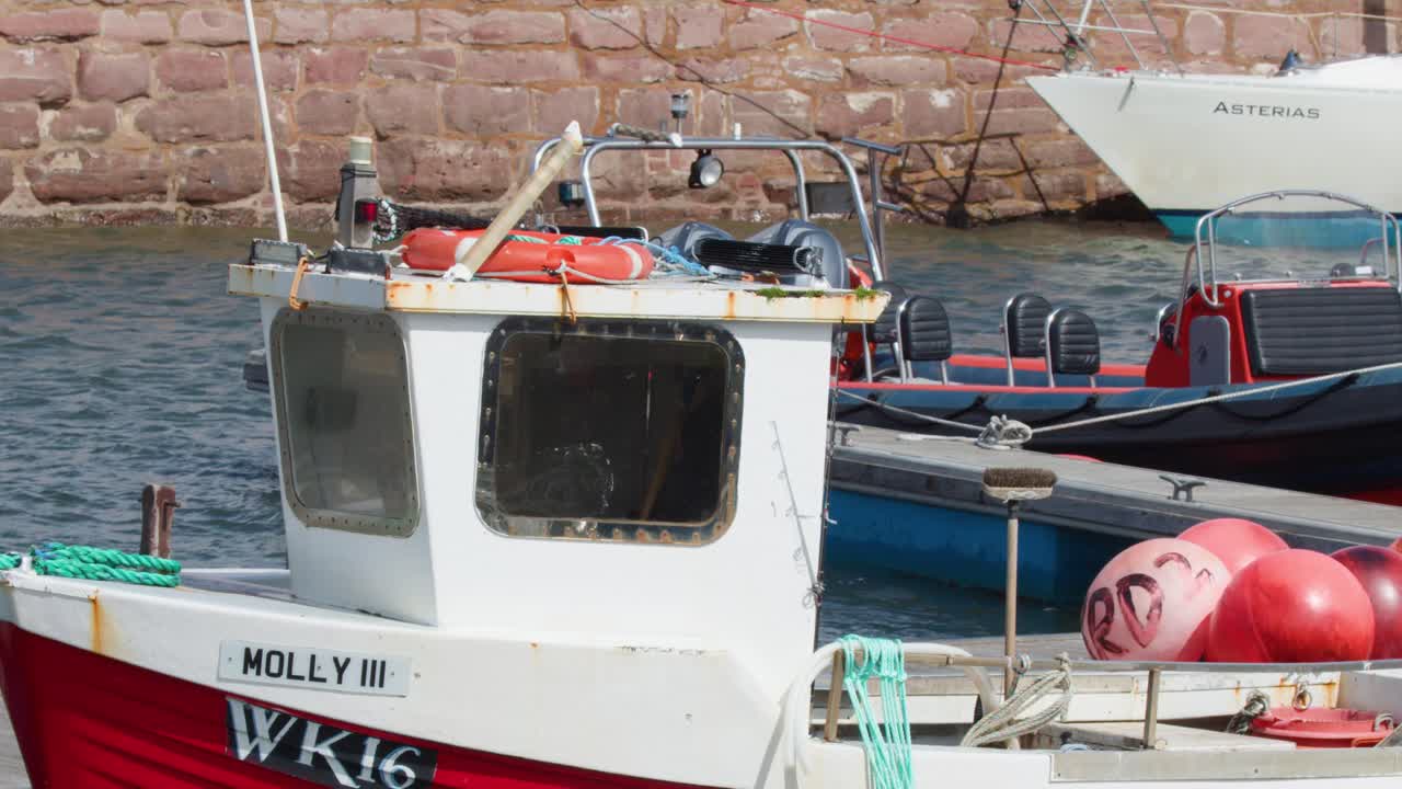 Red fishing boat gently sways at harbor dock, bright daylight, steady camera, marine equipment visible