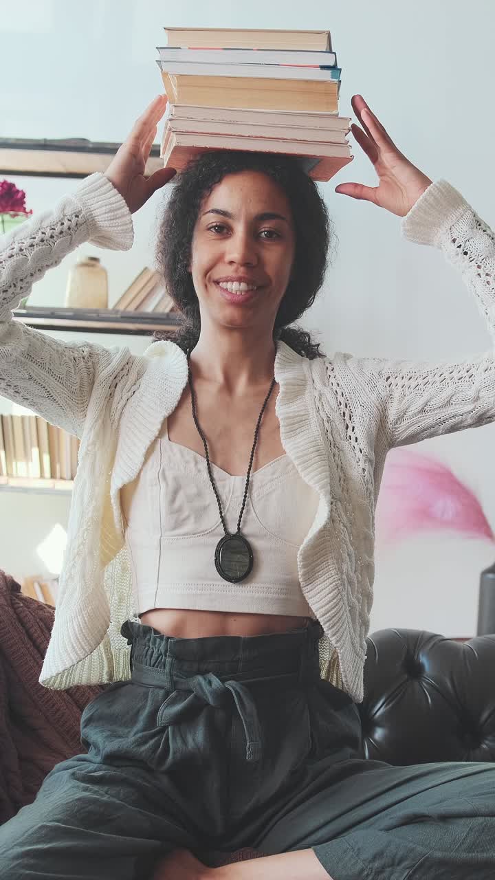 Young optimistic african american woman student puts books on head sits on sofa