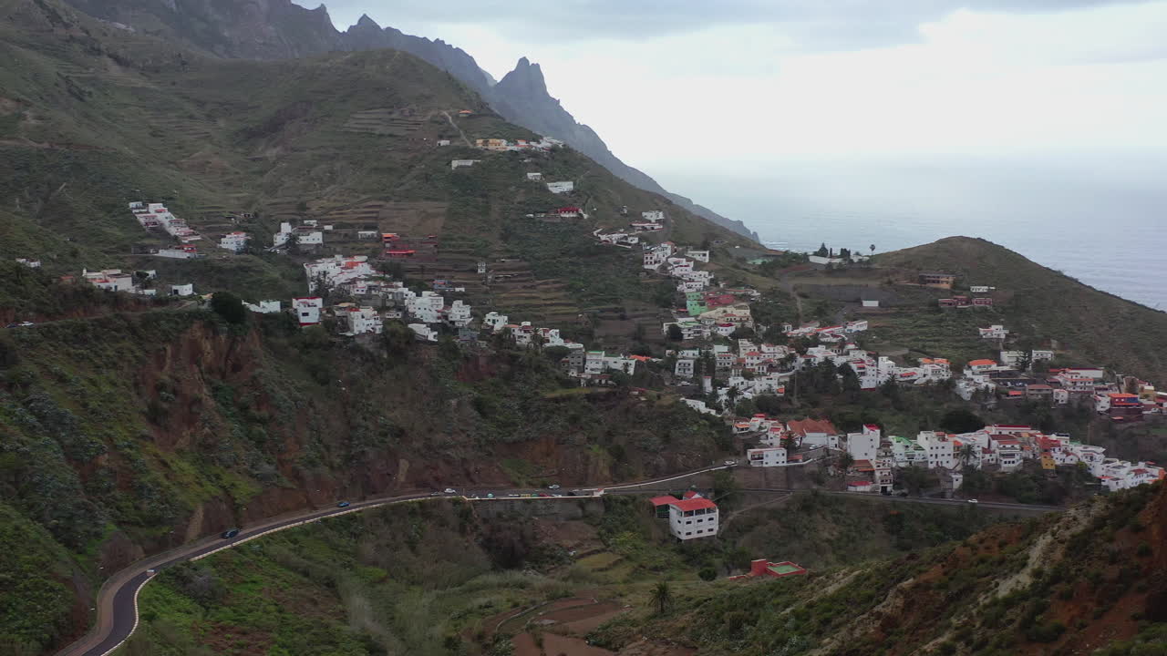 pequeño pueblo de montaña en la empinada ladera de tenerife, islas canarias, españa, picos montañosos agudos arriba, costa del océano atlántico abajo y nubes tormentosas en el horizonte, toma aérea de 4k